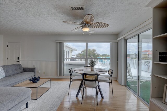 dining space with visible vents, a textured ceiling, a wainscoted wall, a ceiling fan, and light wood-style flooring