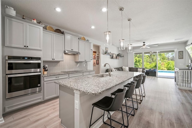 kitchen with a sink, stainless steel double oven, open floor plan, a center island with sink, and under cabinet range hood