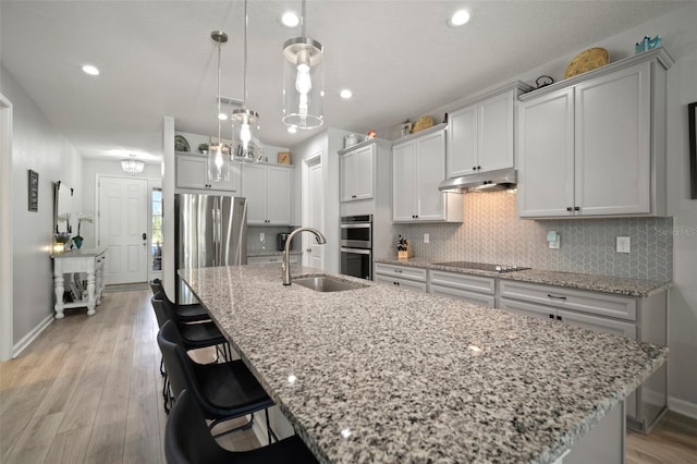 kitchen featuring under cabinet range hood, a sink, backsplash, a center island with sink, and appliances with stainless steel finishes