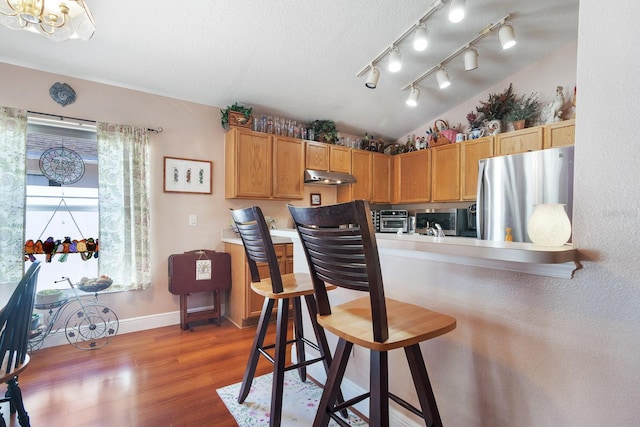 kitchen with wood finished floors, baseboards, a kitchen breakfast bar, under cabinet range hood, and appliances with stainless steel finishes