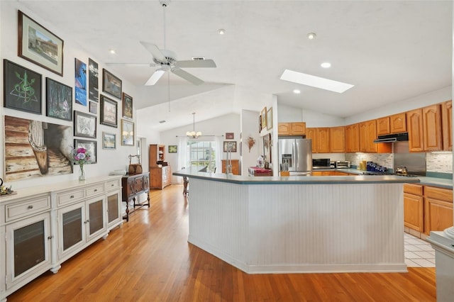 kitchen with stainless steel refrigerator with ice dispenser, under cabinet range hood, vaulted ceiling with skylight, tasteful backsplash, and light wood finished floors