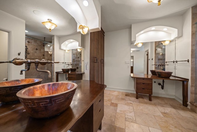 bathroom with baseboards, two vanities, tiled shower, and stone tile flooring
