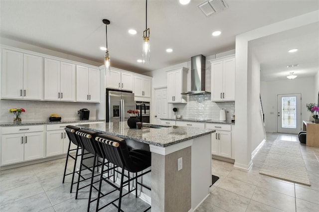 kitchen with appliances with stainless steel finishes, wall chimney range hood, light tile patterned floors, tasteful backsplash, and an island with sink