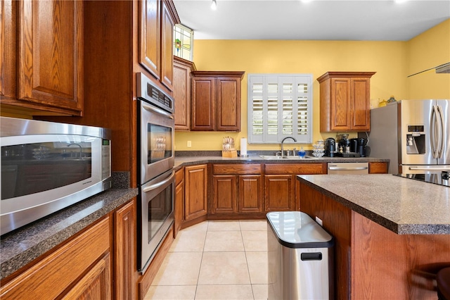 kitchen with appliances with stainless steel finishes, a sink, brown cabinetry, light tile patterned flooring, and dark countertops