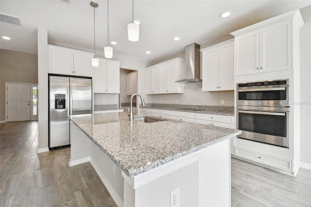 kitchen with wall chimney range hood, appliances with stainless steel finishes, a sink, light wood finished floors, and visible vents
