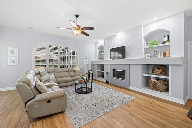 living room featuring built in features, wood finished floors, a fireplace, baseboards, and a ceiling fan