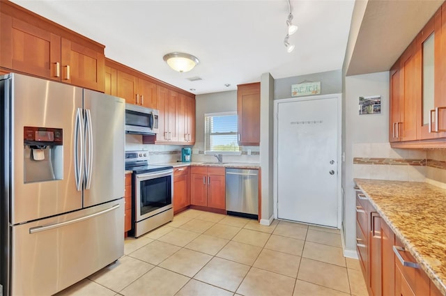 kitchen featuring light tile patterned flooring, light stone countertops, appliances with stainless steel finishes, a sink, and brown cabinetry