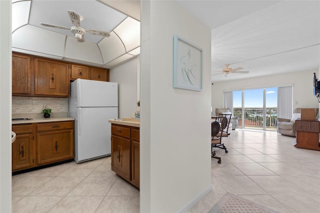 kitchen with brown cabinetry, freestanding refrigerator, backsplash, light countertops, and a ceiling fan