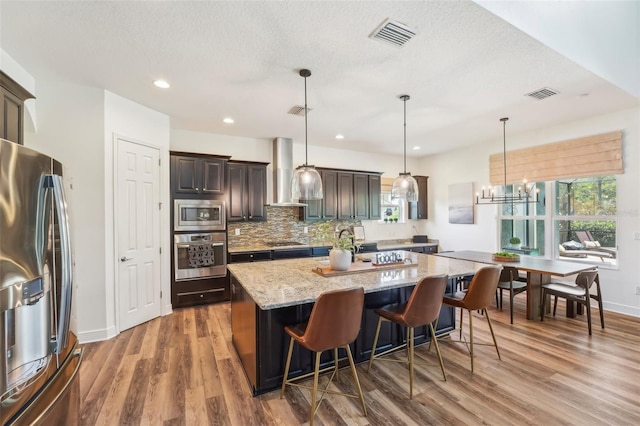 kitchen featuring visible vents, appliances with stainless steel finishes, light stone countertops, a center island with sink, and wall chimney exhaust hood