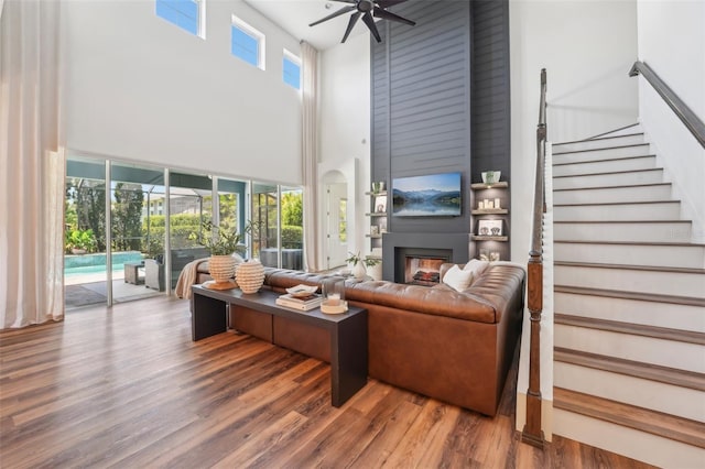 living area with ceiling fan, a fireplace, wood finished floors, a high ceiling, and stairs
