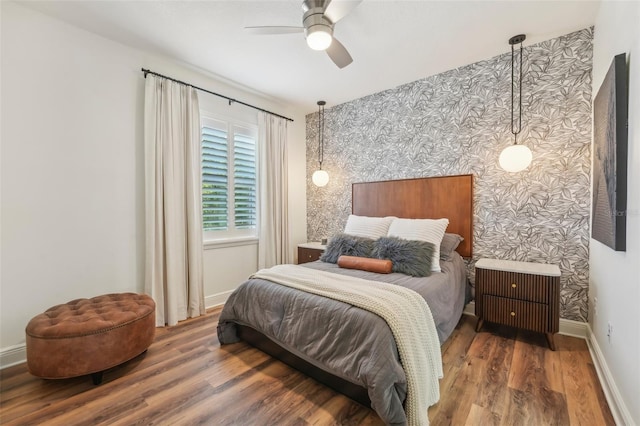 bedroom featuring ceiling fan, wood finished floors, baseboards, an accent wall, and wallpapered walls