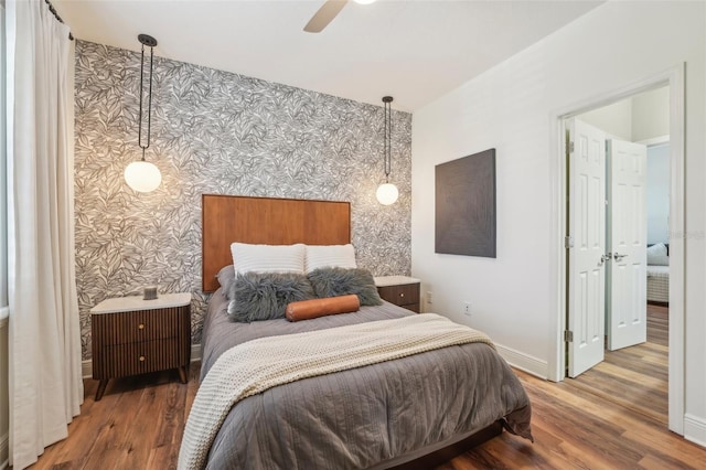 bedroom with wood finished floors, an accent wall, baseboards, wallpapered walls, and a ceiling fan