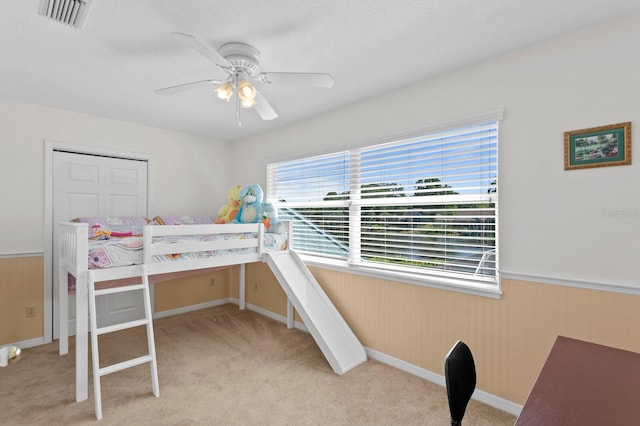 carpeted bedroom with wooden walls, a ceiling fan, visible vents, and a wainscoted wall