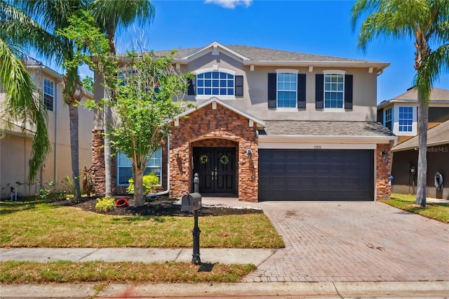traditional-style house featuring a garage, stone siding, a front yard, decorative driveway, and stucco siding