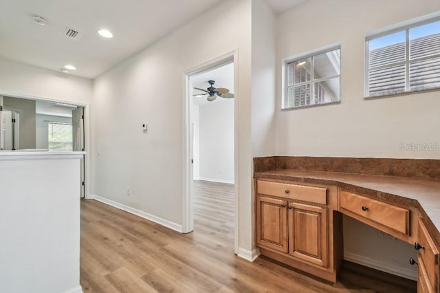 interior space featuring ceiling fan, built in desk, light wood-style flooring, baseboards, and visible vents