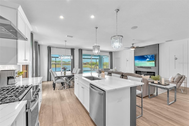 kitchen featuring light wood-style floors, wall chimney range hood, a sink, ceiling fan with notable chandelier, and stainless steel appliances