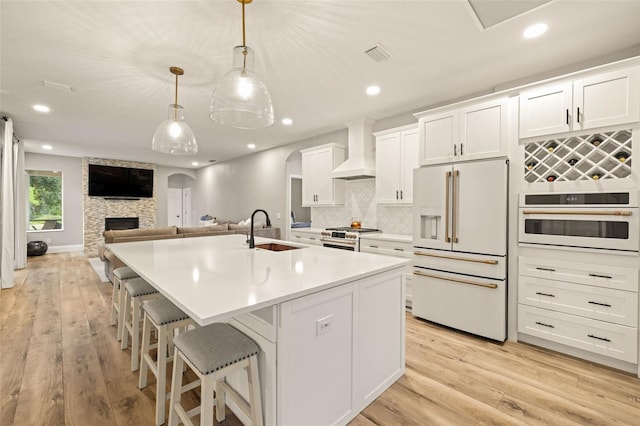 kitchen with a kitchen island with sink, high end appliances, light wood-type flooring, wall chimney exhaust hood, and a sink