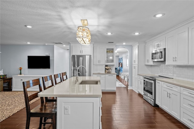 kitchen featuring a sink, a kitchen island with sink, arched walkways, stainless steel appliances, and dark wood-style floors