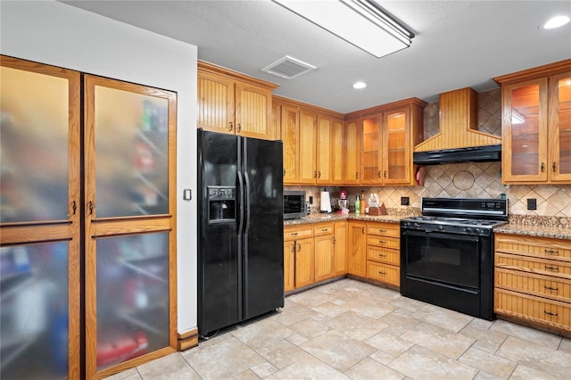 kitchen with black appliances, premium range hood, tasteful backsplash, light stone countertops, and glass insert cabinets