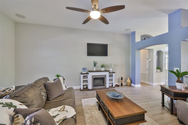 living area with ceiling fan, light wood-style floors, arched walkways, a glass covered fireplace, and baseboards