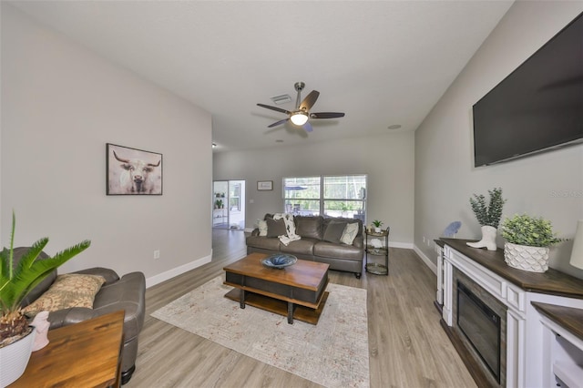 living area featuring baseboards, ceiling fan, a glass covered fireplace, and light wood-style flooring