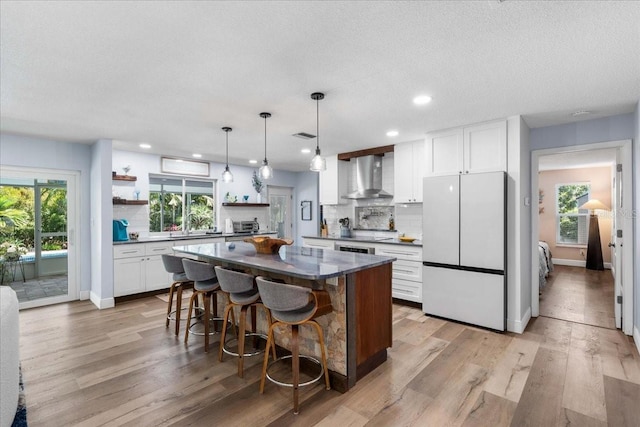 kitchen featuring freestanding refrigerator, wall chimney range hood, open shelves, dark countertops, and healthy amount of natural light