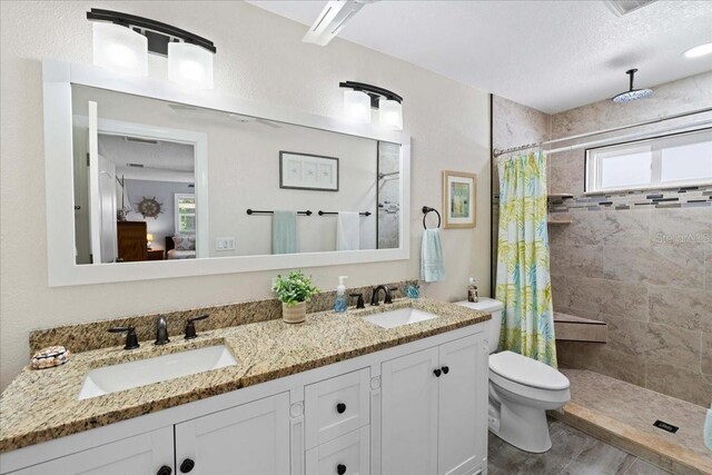 ensuite bathroom featuring a tile shower, toilet, wood finished floors, double vanity, and a textured ceiling
