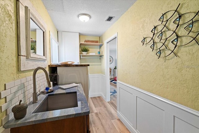 kitchen featuring a textured ceiling, a textured wall, light wood finished floors, a sink, and a wainscoted wall