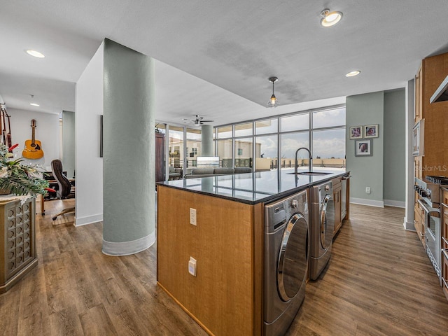 kitchen with a sink, brown cabinets, baseboards, a center island with sink, and dark wood-style flooring