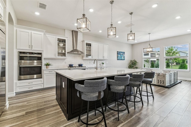 kitchen with wall chimney exhaust hood, light countertops, light wood finished floors, recessed lighting, and a kitchen island with sink