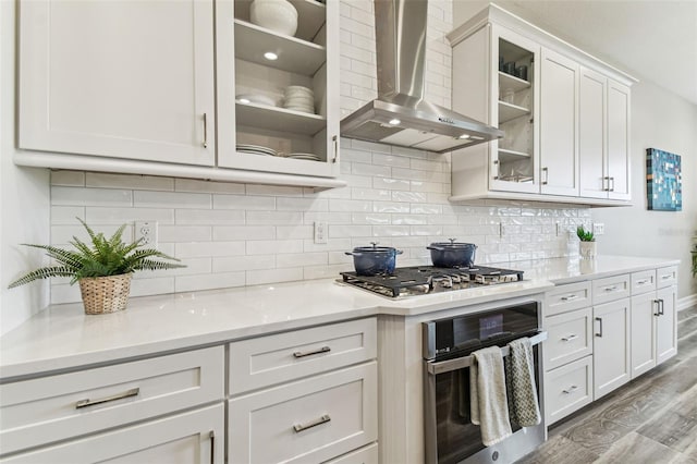 kitchen with appliances with stainless steel finishes, wall chimney exhaust hood, white cabinets, light wood-type flooring, and light countertops