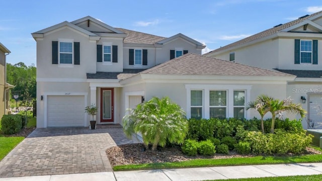 traditional-style house with an attached garage, decorative driveway, roof with shingles, and stucco siding
