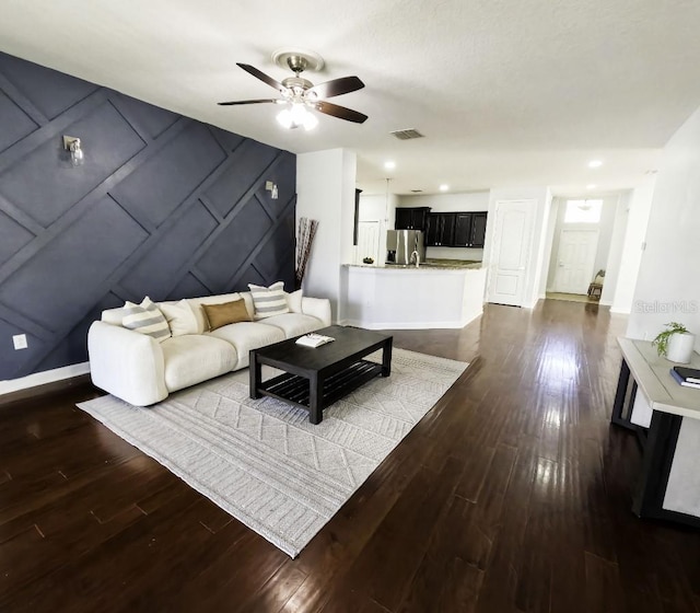 living area featuring visible vents, a decorative wall, baseboards, ceiling fan, and dark wood finished floors