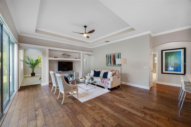 living area featuring ornamental molding, dark wood finished floors, ceiling fan, arched walkways, and a tray ceiling