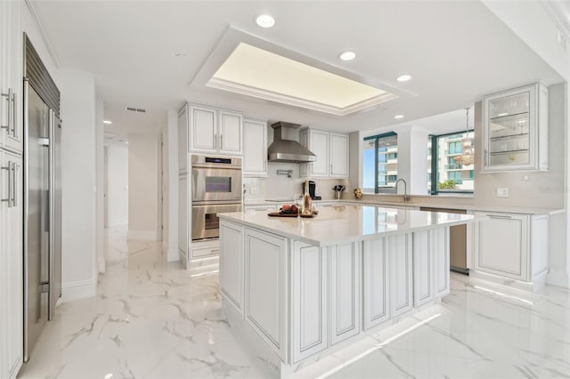 kitchen with marble finish floor, light countertops, wall chimney exhaust hood, a tray ceiling, and stainless steel appliances