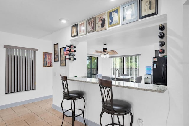 kitchen featuring a breakfast bar, freestanding refrigerator, light stone counters, ceiling fan, and baseboards