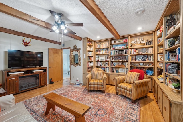 living area featuring ceiling fan, a textured ceiling, light wood-type flooring, beamed ceiling, and a glass covered fireplace