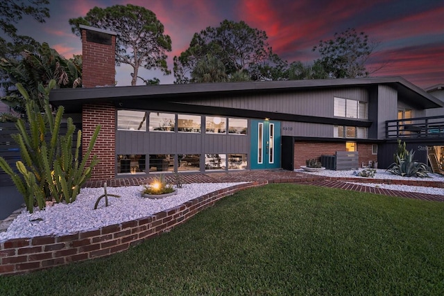 back of house at dusk with central AC, brick siding, a chimney, a yard, and a fire pit