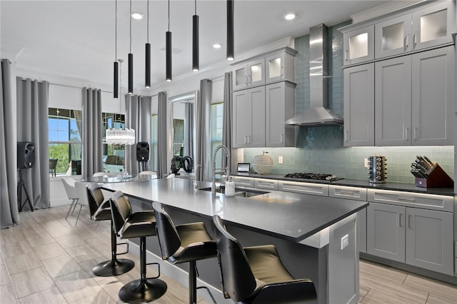 kitchen with wall chimney range hood, tasteful backsplash, a center island with sink, a sink, and gray cabinetry