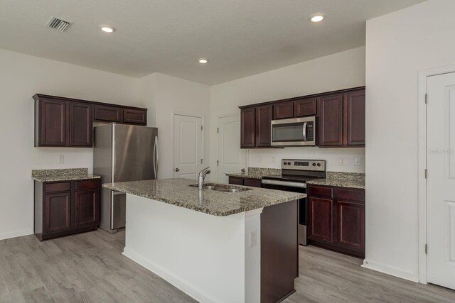 kitchen with stainless steel appliances, a sink, stone countertops, light wood-style floors, and an island with sink