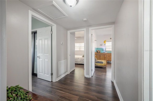 hall featuring dark wood-style flooring, baseboards, visible vents, and attic access