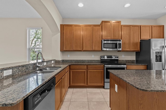 kitchen featuring appliances with stainless steel finishes, a sink, brown cabinetry, dark stone countertops, and recessed lighting