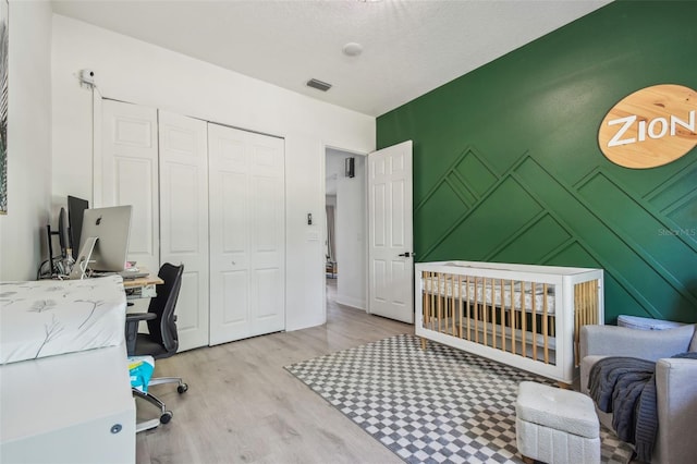 bedroom with a nursery area, a decorative wall, wood finished floors, visible vents, and a closet