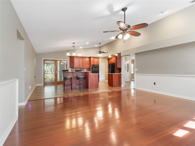 unfurnished living room featuring lofted ceiling, light wood finished floors, wainscoting, and ceiling fan with notable chandelier