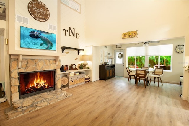 living area featuring a towering ceiling, a ceiling fan, wood finished floors, a stone fireplace, and baseboards