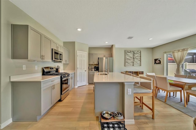 kitchen with appliances with stainless steel finishes, light wood finished floors, a kitchen island with sink, a kitchen breakfast bar, and gray cabinets