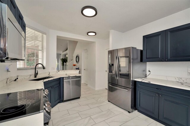 kitchen featuring appliances with stainless steel finishes, a sink, light stone counters, blue cabinetry, and baseboards