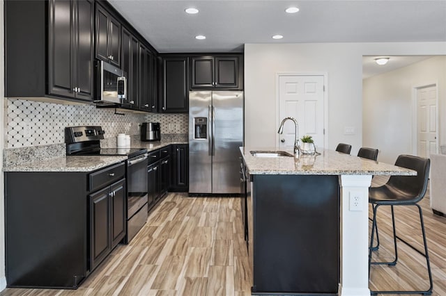 kitchen featuring stainless steel appliances, a sink, dark cabinetry, a kitchen breakfast bar, and an island with sink