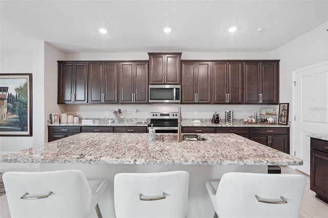 kitchen featuring stainless steel appliances, a breakfast bar, dark brown cabinetry, light stone countertops, and recessed lighting