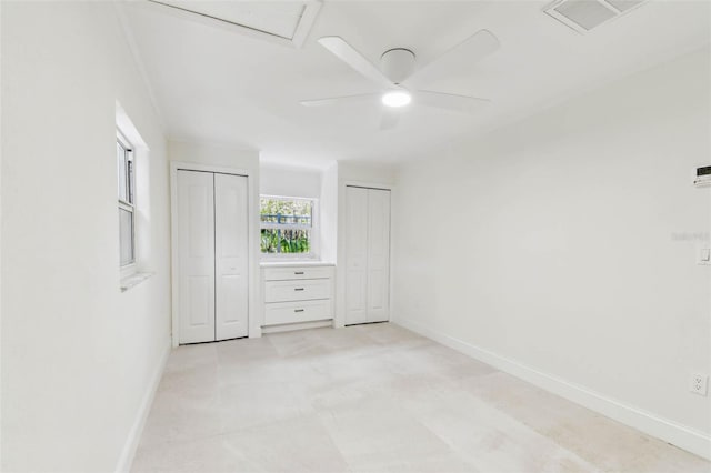 unfurnished bedroom featuring two closets, baseboards, ceiling fan, and attic access